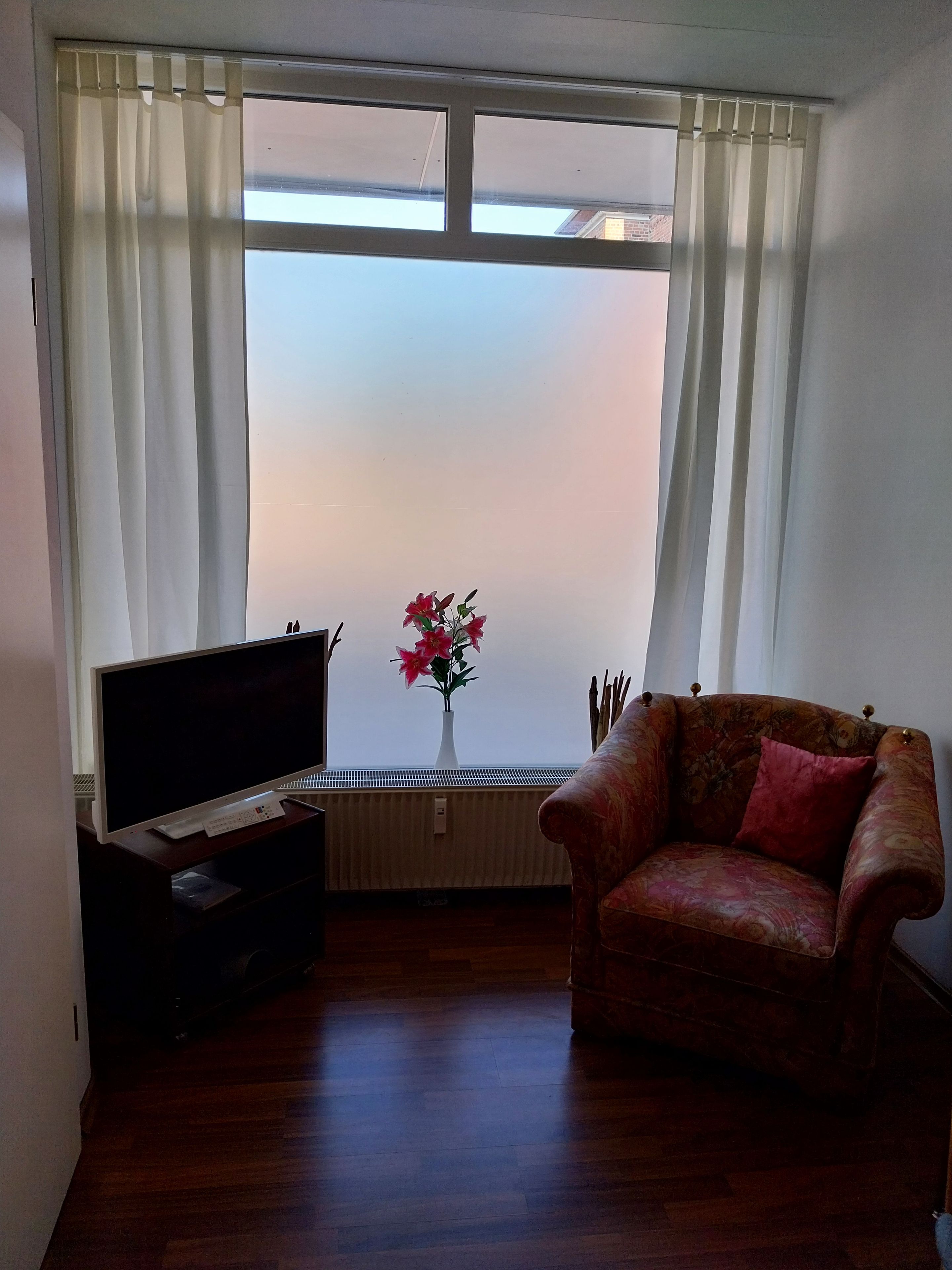 Ett vardagsrum med en soffa, en tv, ett element och ett fönster med gardiner.