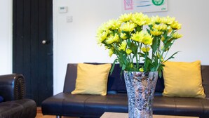 Un salon avec une porte noire, un canapé avec des coussins jaunes et un vase transparent contenant des fleurs jaunes, posé sur une table.