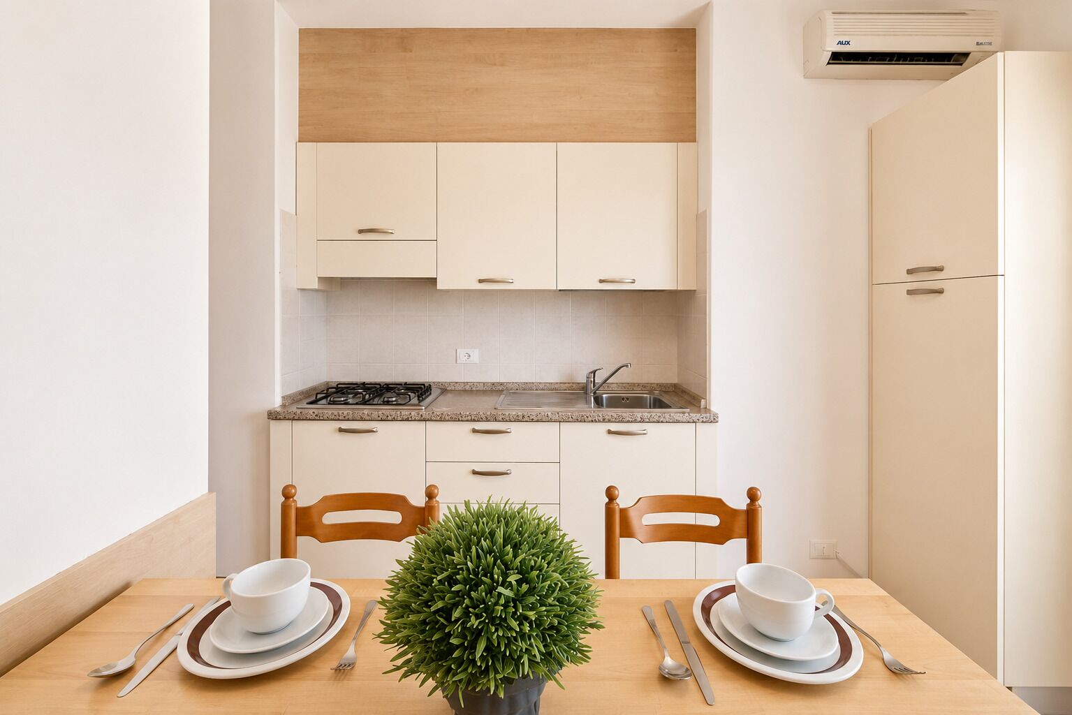 Une cuisine de taille réduite avec des meubles blancs, une cuisinière et un évier. On y trouve une table à manger avec des chaises et une plante en pot.