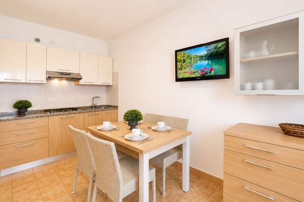 A modern kitchen with white cabinets, a dining table with chairs, a TV mounted on the wall, and a small cabinet with a basket. - Residence Columbia (San Michele al Tagliamento)