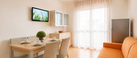 A modern living room with a dining area, a television mounted on the wall, a refrigerator, and a sofa.