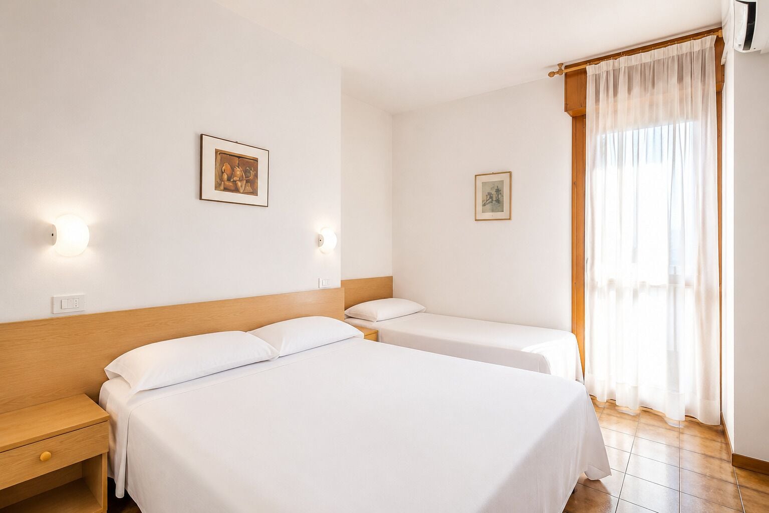 A hotel room with two beds, a wooden headboard, a bedside table, a nightstand, a window with sheer curtains, and two wall-mounted light fixtures.
