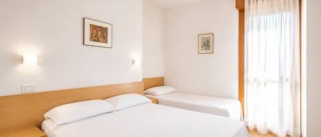 A hotel room with two beds, a wooden headboard, a bedside table, a nightstand, a window with sheer curtains, and two wall-mounted light fixtures.