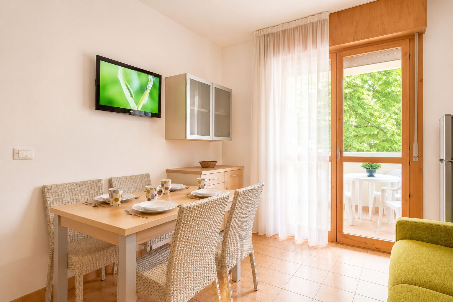 A dining area with a table set for four, a television mounted above, and a sliding glass door leading to an outdoor seating area.