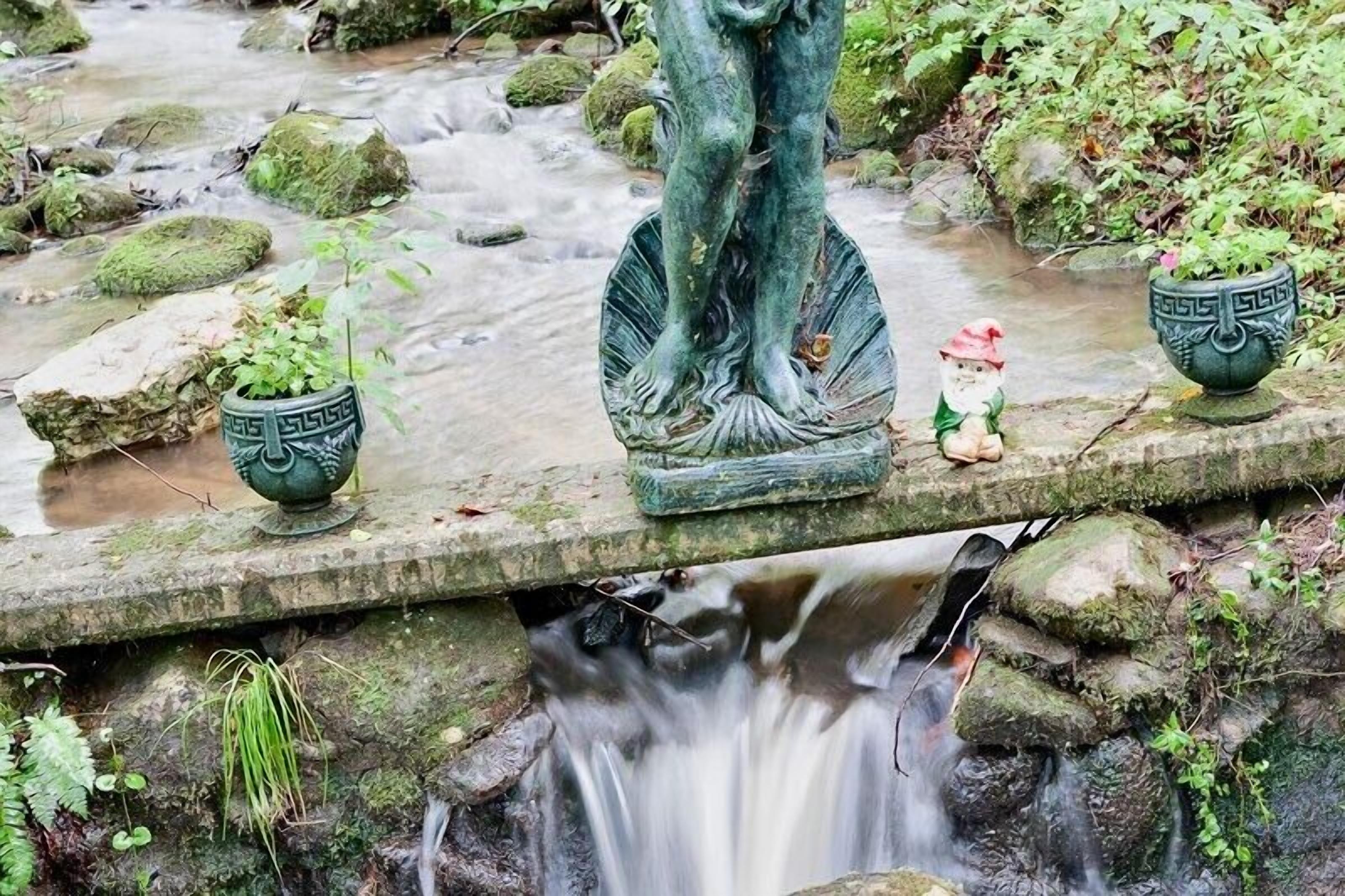 Un puente de piedra sobre un arroyo con una estatua de una figura y dos macetas decorativas.