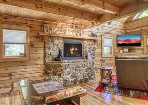 A cozy cabin interior with a stone fireplace, wooden walls, a television, and a glass table with a menu. - Romantic 1BR Couples Cabin: Hot Tub, Mountain Views, Arcade - Near Dollywood (Sevierville)