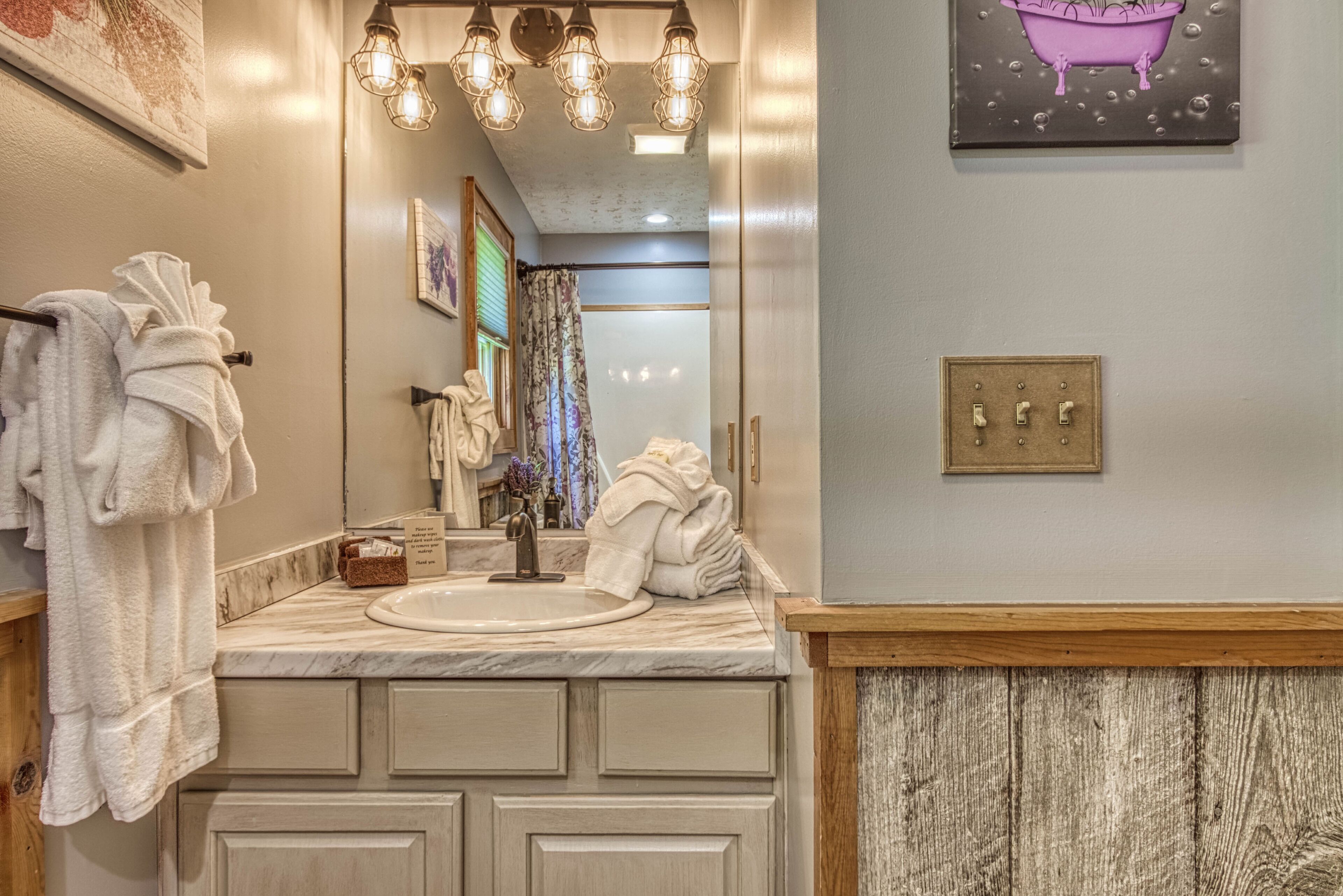 Une salle de bain avec un lavabo, un miroir et un porte-serviettes. Il y a un luminaire au-dessus du miroir et un tableau accroché au mur.