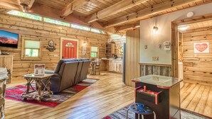Interior de una cabaña de troncos con sala de estar, cocina y una mesa de juegos.