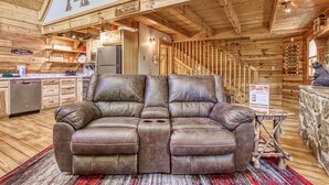 Un interior de cabaña acogedor con sofá de cuero, paredes de madera, una zona de cocina y una escalera que sube a un altillo.