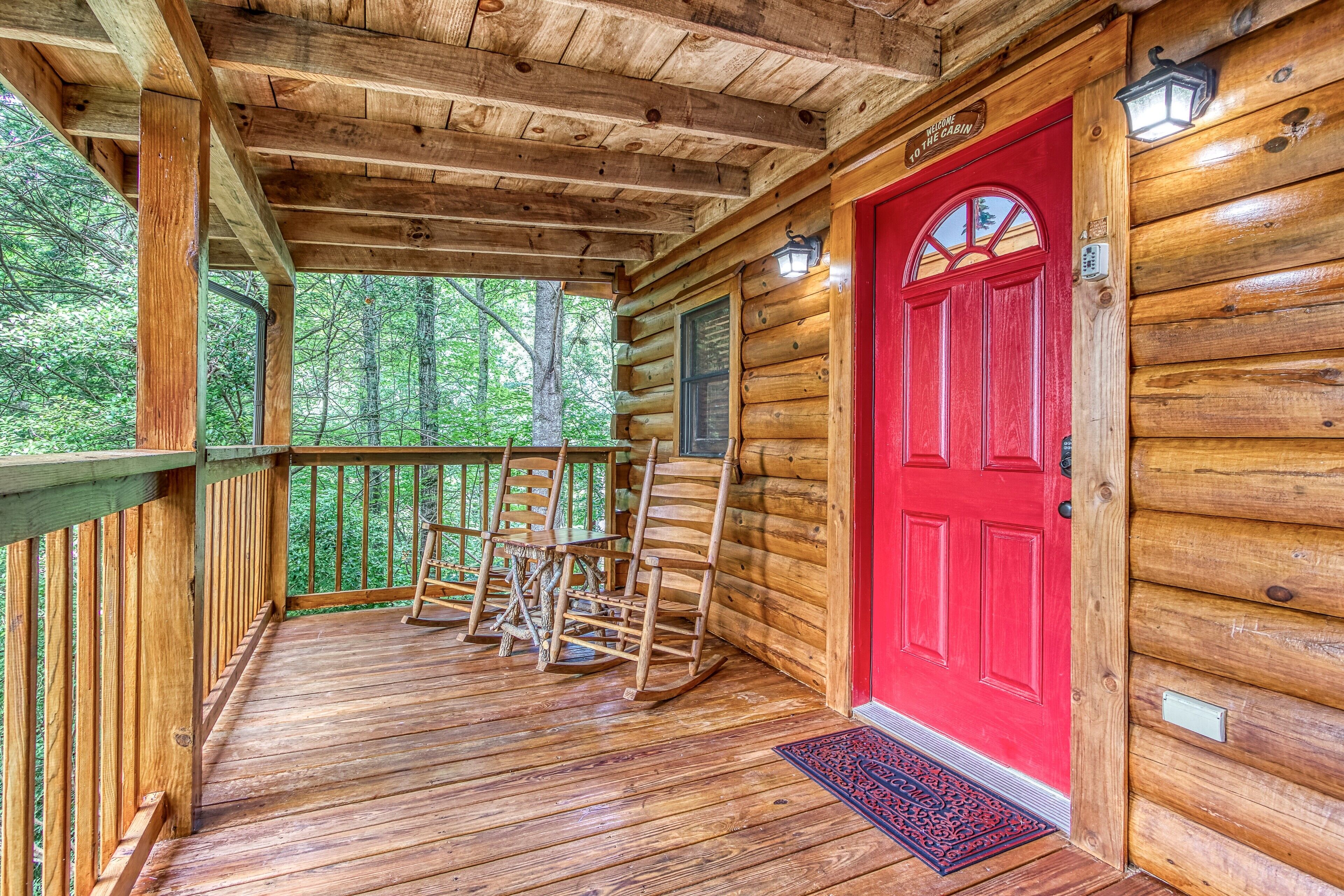 A wooden cabin with a red door, a porch with rocking chairs, and a view of the forest.