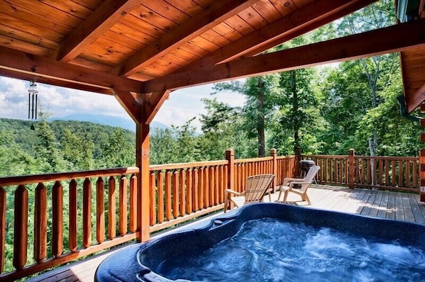 A wooden deck with a hot tub, chairs, and a view of a forested mountain.