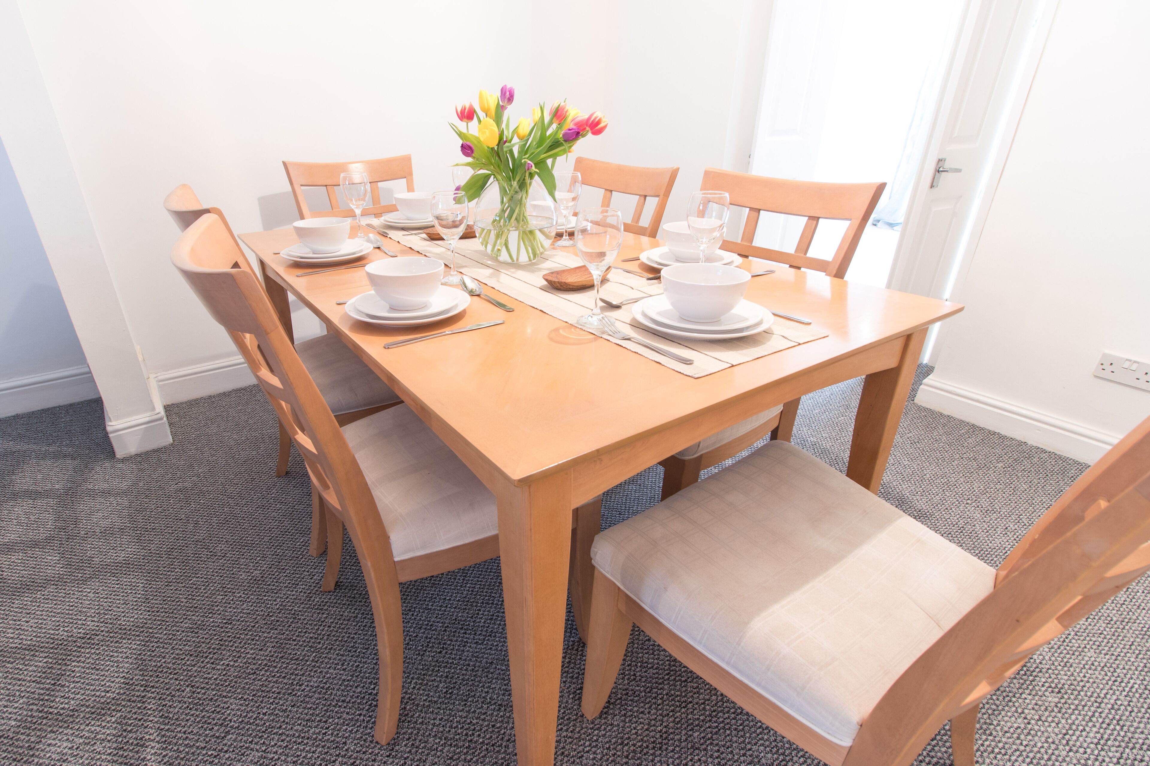 A dining table set with white plates, bowls, and cutlery, surrounded by wooden chairs with cushioned seats.