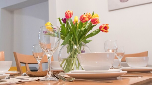Uma mesa de jantar posta com pratos, tigelas e talheres brancos, acompanhada de um vaso de tulipas.