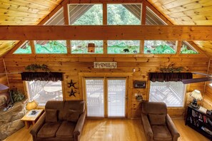 A cozy cabin interior with a high ceiling, wooden walls, and a large window overlooking a forest.