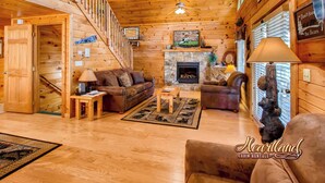 A cabin interior with a fireplace, wooden walls, a staircase, and a cozy seating area.