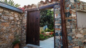 A stone wall entrance with a wooden door, a potted plant, and a garden with flowers. - Estia Anogaia Cottage (Ora)