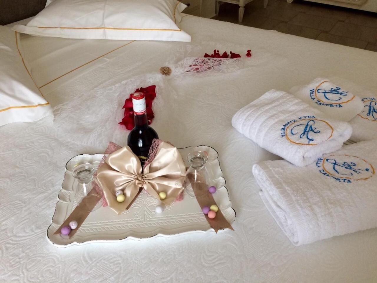 A neatly arranged bed with a decorative tray, a bottle of wine, and towels with embroidered logos.