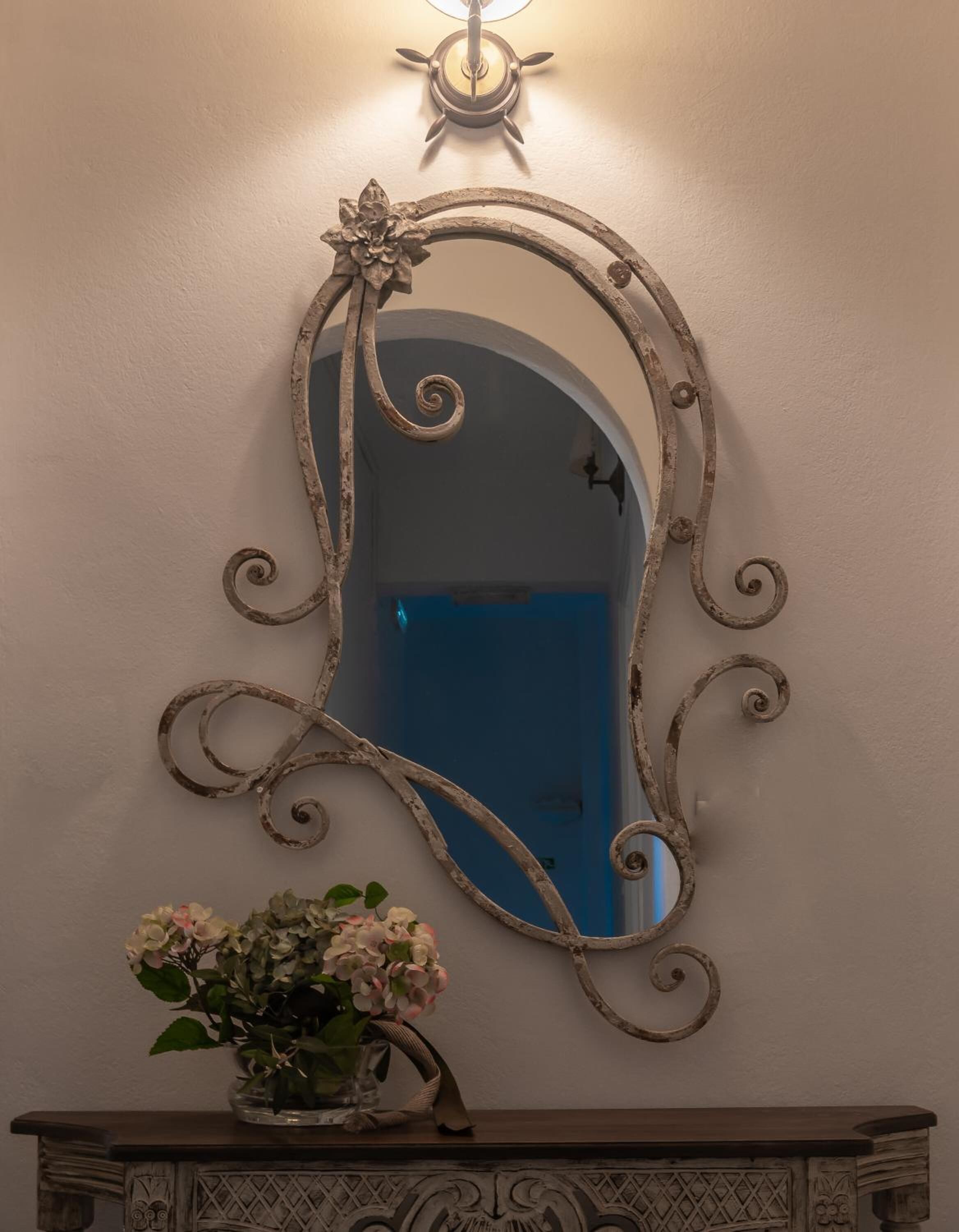 A mirror with ornate metalwork and a floral design, mounted on a wall above a wooden console table with a vase of flowers.