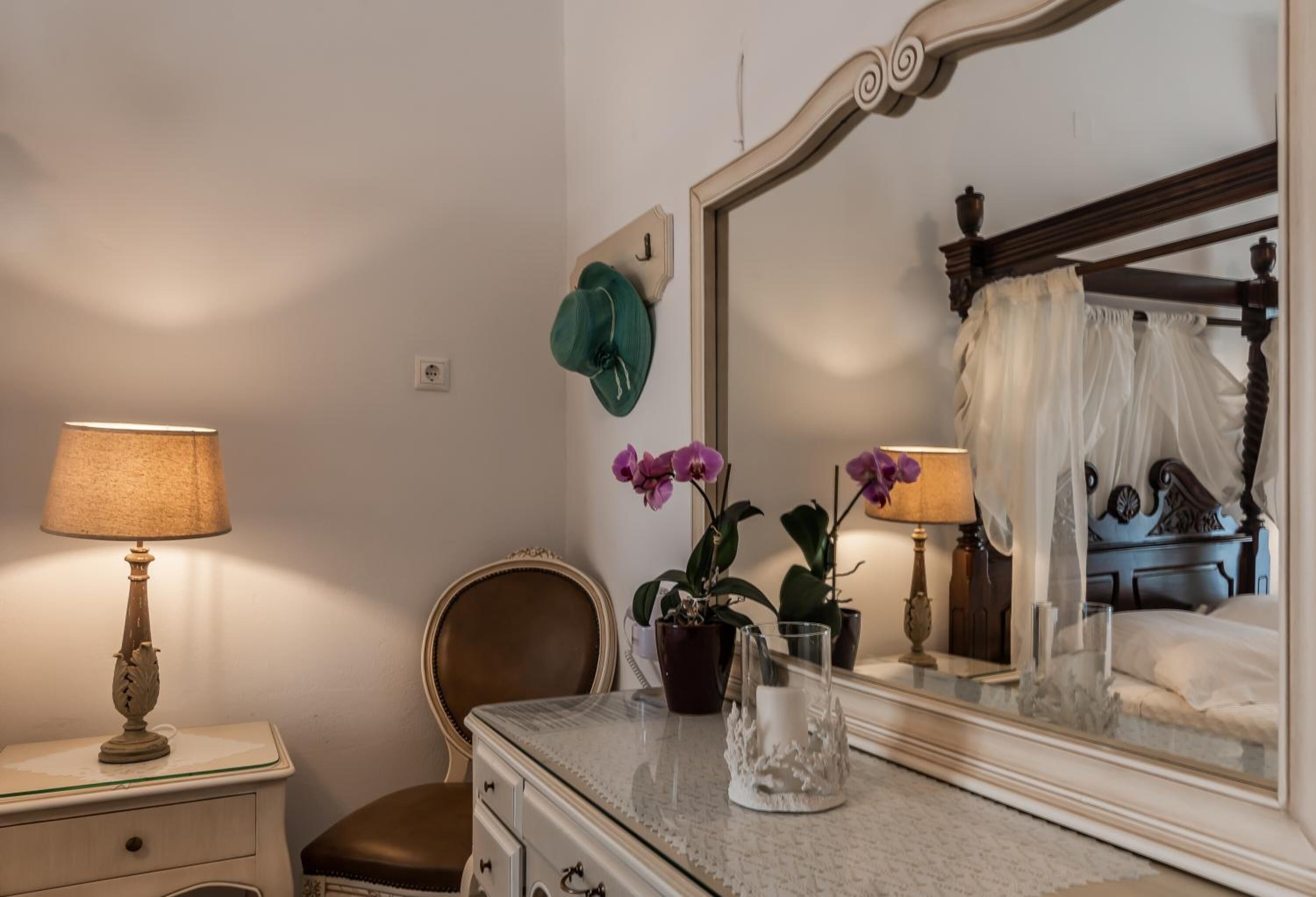 A bedroom with a bed, a dresser, a chair, a nightstand with a lamp, and a mirror reflecting the bed and a window.
