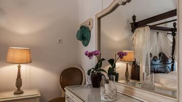 A bedroom with a bed, a dresser, a chair, a nightstand with a lamp, and a mirror reflecting the bed and a window.