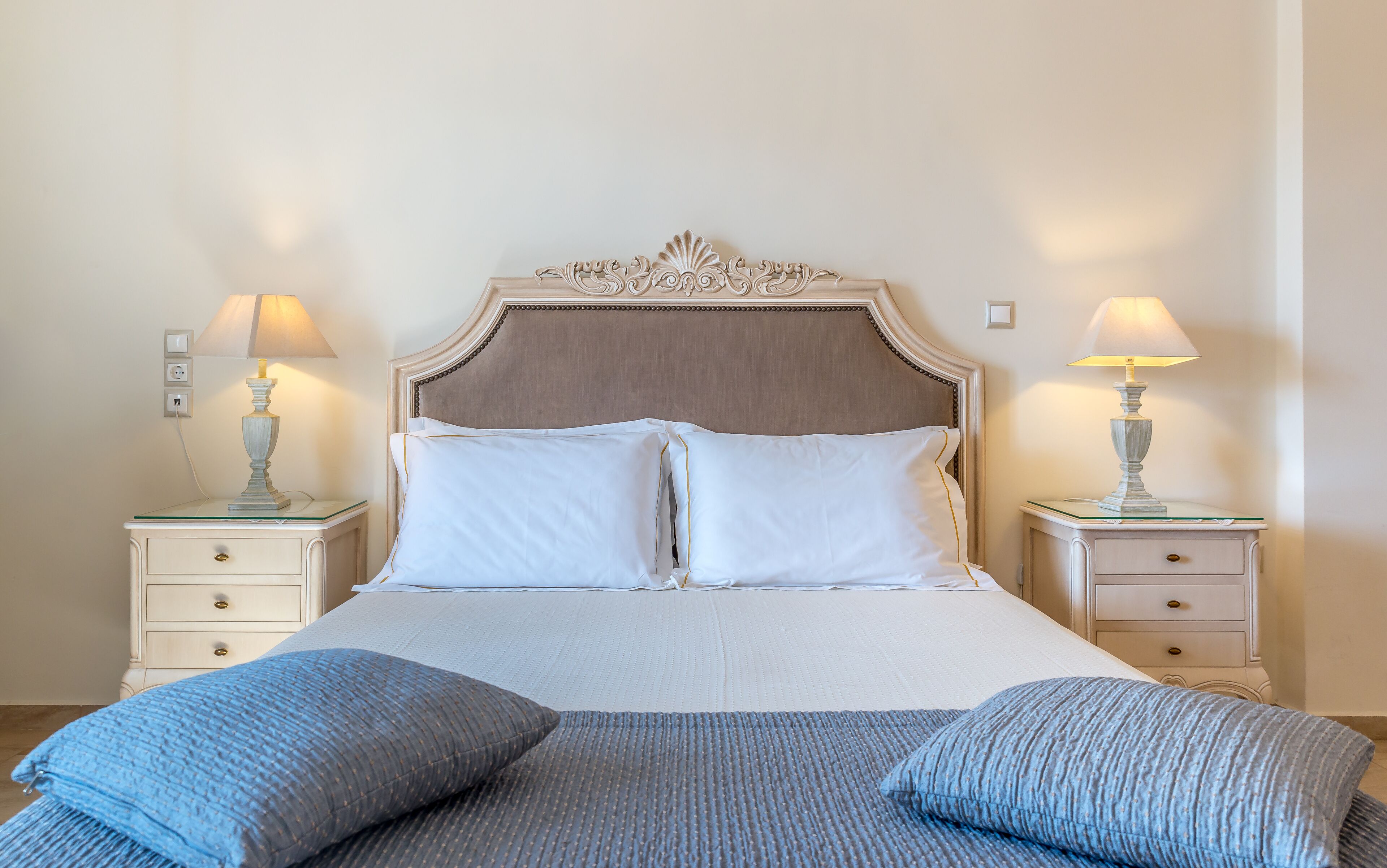 A neatly made bed with white linens and two blue pillows, flanked by two bedside tables with lamps.