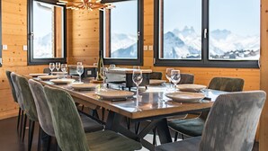 Une salle à manger avec une longue table dressée pour six personnes, des murs lambrés, de grandes fenêtres offrant une vue sur les montagnes et un lustre moderne.