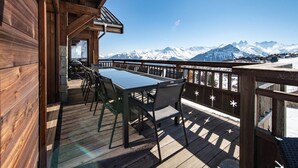 Une terrasse en bois avec une table et des chaises, offrant une vue sur une chaîne de montagnes enneigées.