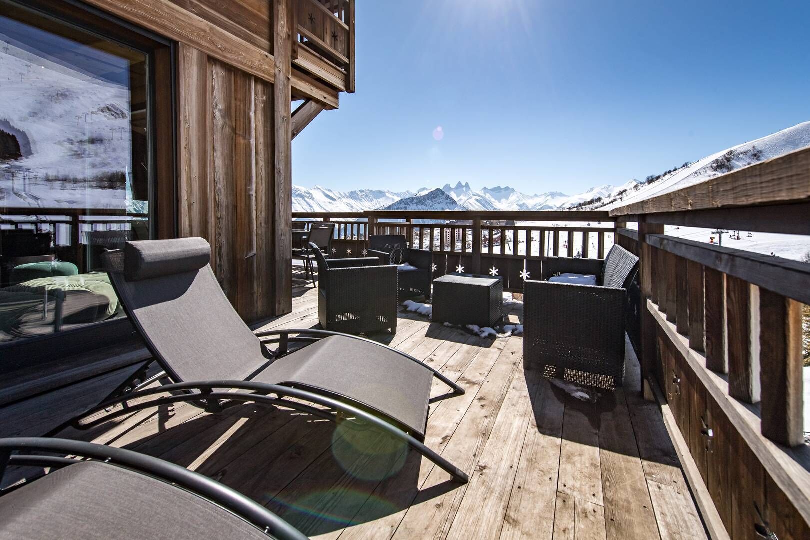 A wooden deck with lounge chairs and tables, overlooking a snowy mountain range.