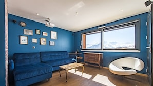 Un salon avec un canapé bleu, une table basse en bois, une chaise blanche, une commode en bois et une grande fenêtre donnant sur une vue sur les montagnes.