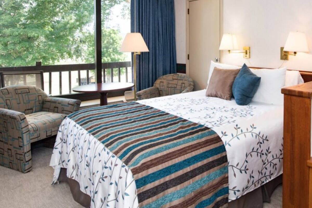 Habitación de hotel con cama, dos sillas, una mesita y balcón con vista a árboles.