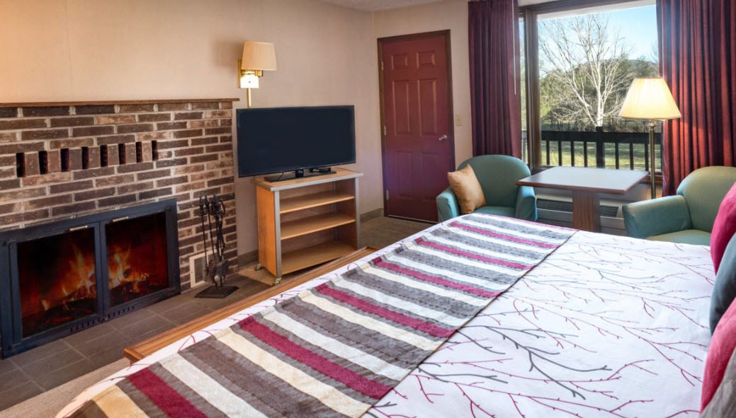 Ein Hotelzimmer mit Kamin aus Ziegelsteinen, einem Fernseher auf einem Holzboden, einem Bett mit gestreifter Bettwäsche, einem Fenster mit Vorhängen und Blick auf Bäume.