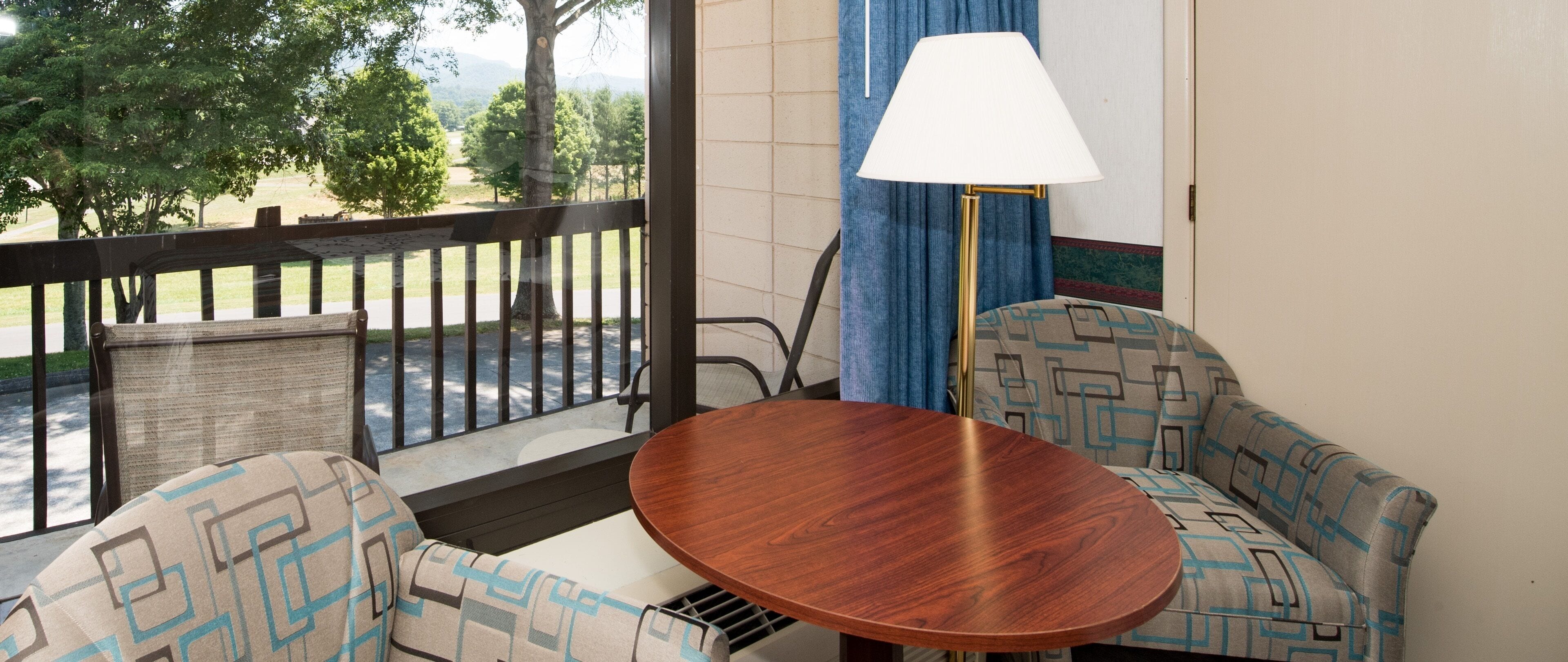 Een balkon met een houten tafel en twee gestreept beklede banken, een lamp, en uitzicht op bomen en een weg.