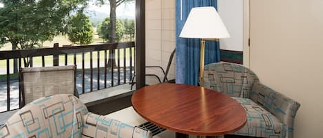 A balcony with a wooden table and two patterned sofas, a lamp, and a view of trees and a road.