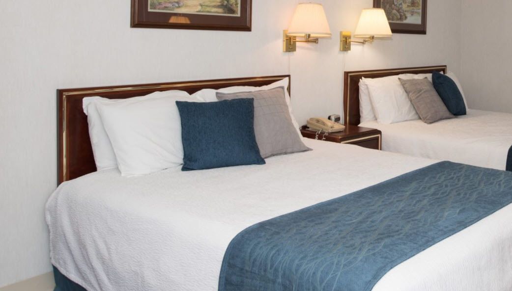 Two double beds with white and blue bedding, wooden headboards, bedside tables with lamps, and a framed picture on the wall.