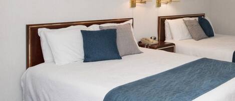Two double beds with white and blue bedding, wooden headboards, bedside tables with lamps, and a framed picture on the wall.