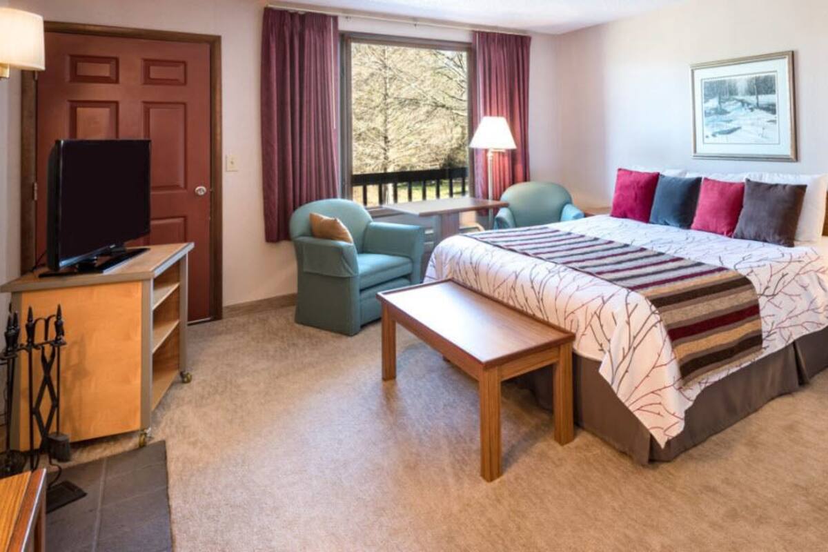 Una habitación de hotel con cama, escritorio, televisión, chimenea y ventana con cortinas.