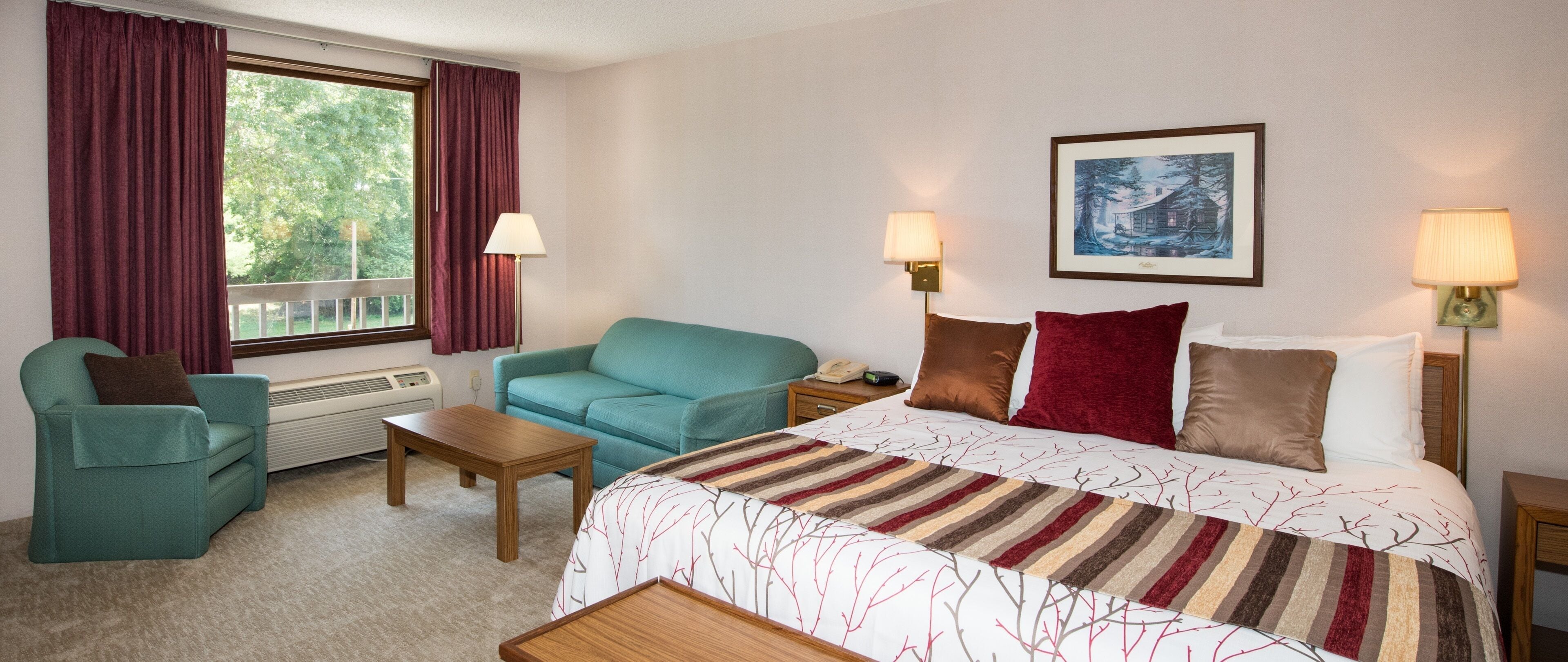 Una habitación de hotel con una cama, un sillón verde azulado, una mesita de noche de madera, una pequeña mesa y dos lámparas fijadas a la pared.