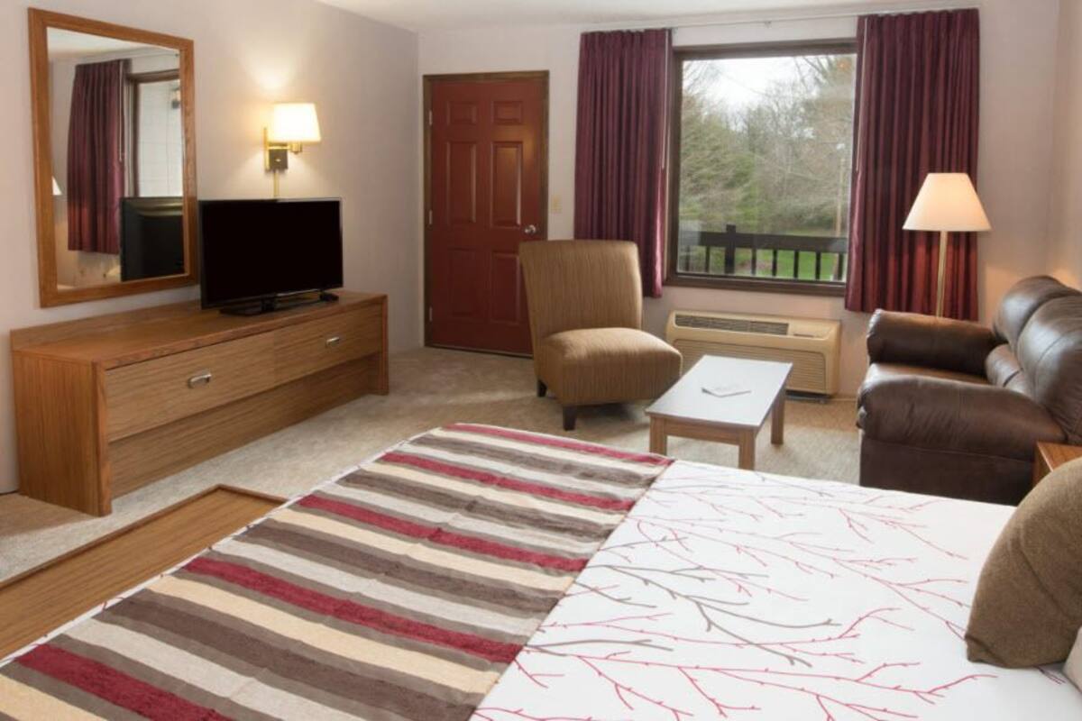 Una habitación de hotel con cama, televisor sobre una cómoda de madera, una silla, un sofá, una mesita y una ventana con cortinas.