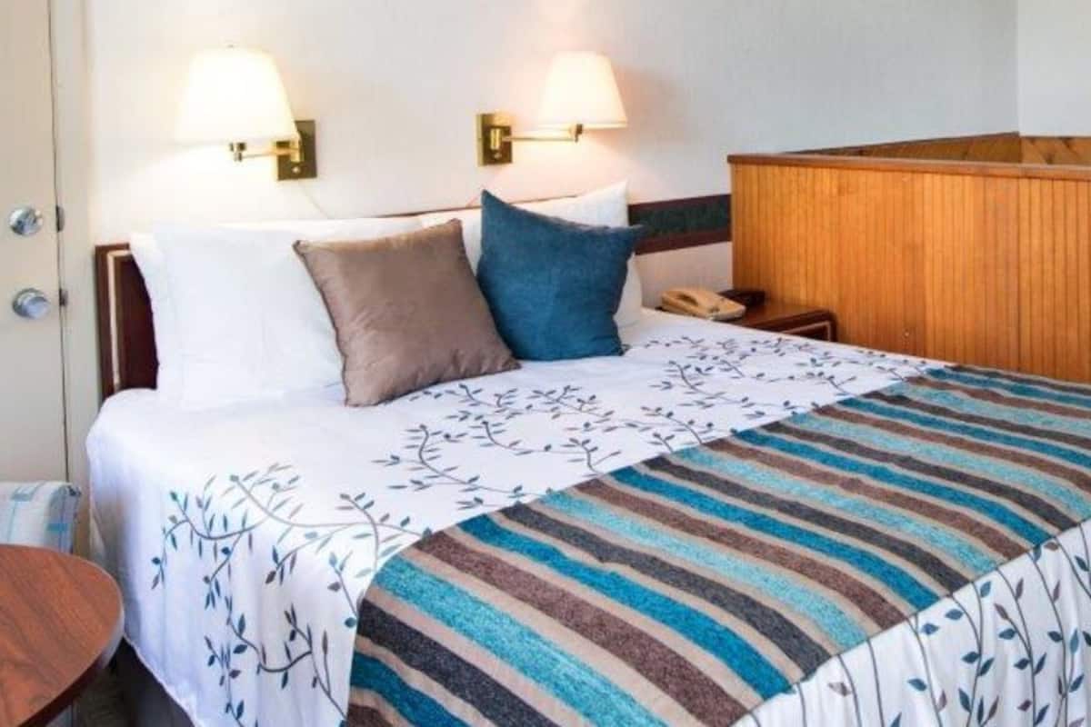 Habitación de hotel con una cama, dos lámparas fijadas a la pared, un cabecero de madera, una mesita de noche y un pequeño mueble de madera.