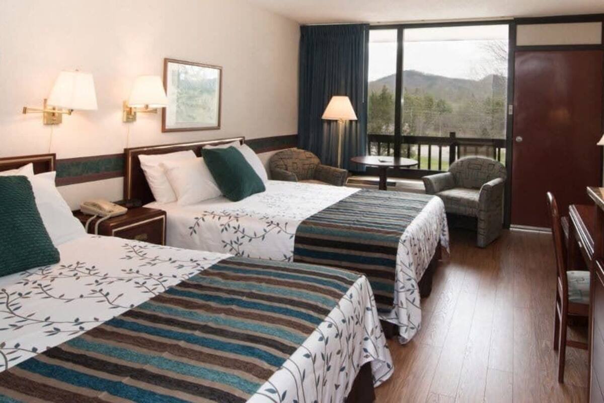 Habitación de hotel con dos camas, un escritorio, una silla y vista a las montañas a través del ventanal.