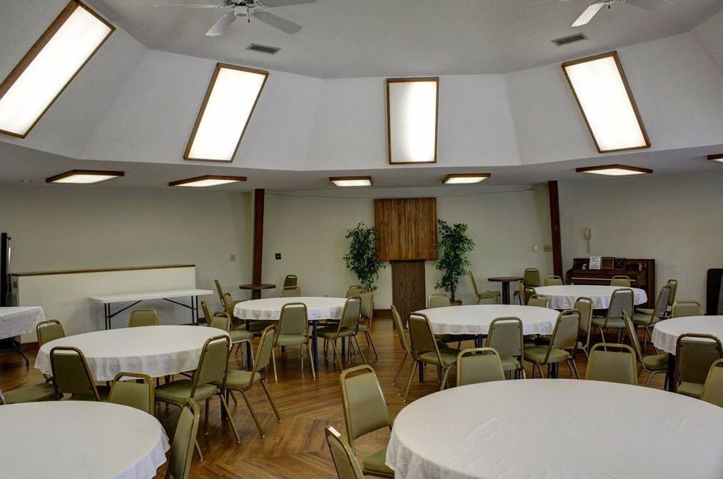 A conference room with multiple round tables and chairs, ceiling lights, and a wooden panel on the wall.