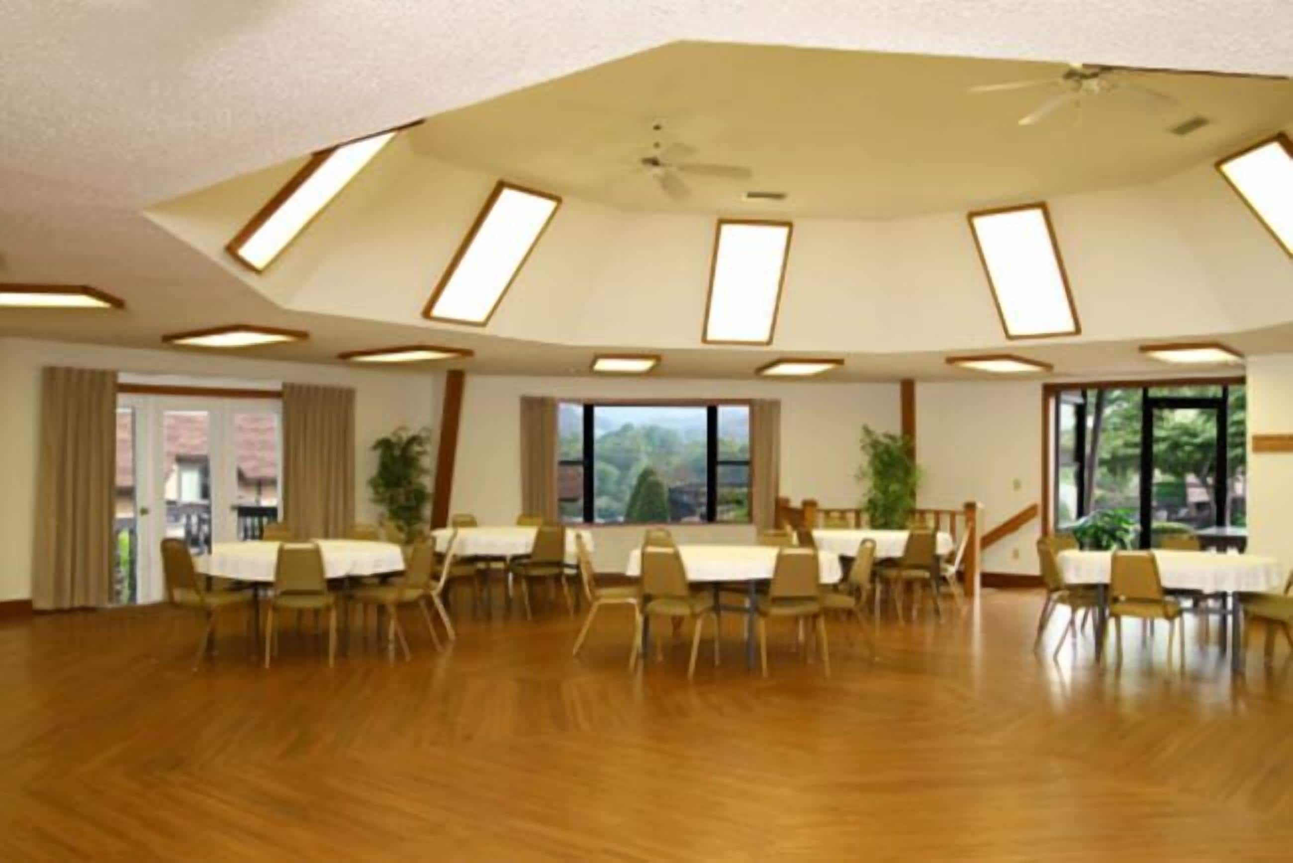 Une pièce spacieuse avec un plafond haut, des fenêtres de toit et un ventilateur de plafond. Des tables et des chaises sont disposées pour les repas.