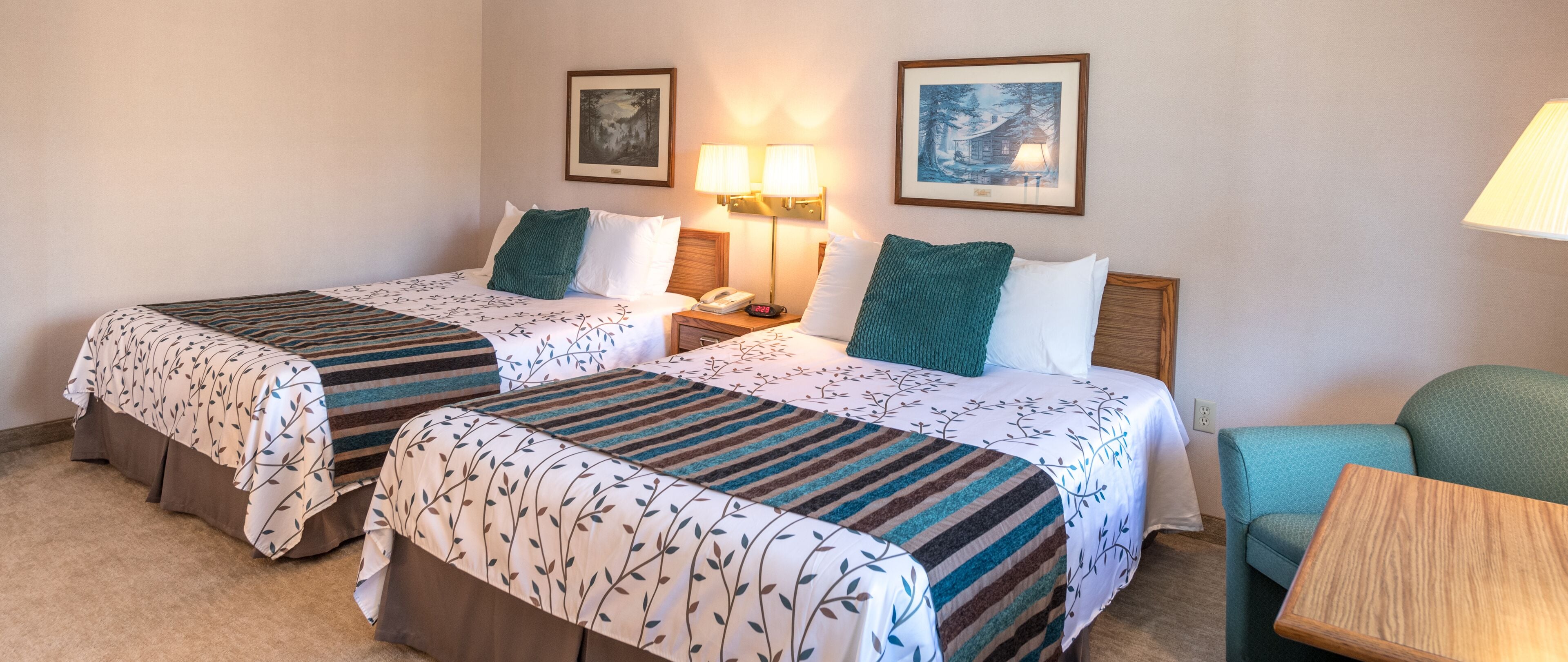 Een hotelkamer met twee bedden, elk met een gestreept beddekleed, een nachtkastje met een lamp, en twee ingelijste foto’s aan de muur.