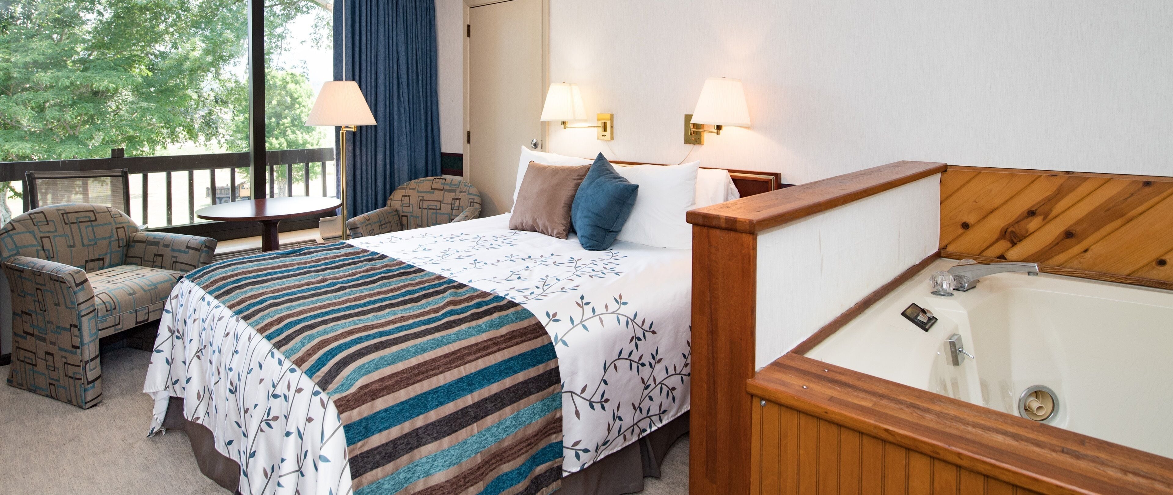 Ein Hotelzimmer mit einem Bett, einem Holzkopfteil, einer Badewanne, einem Sessel, einem Tisch und Blick auf Bäume durch das Fenster.