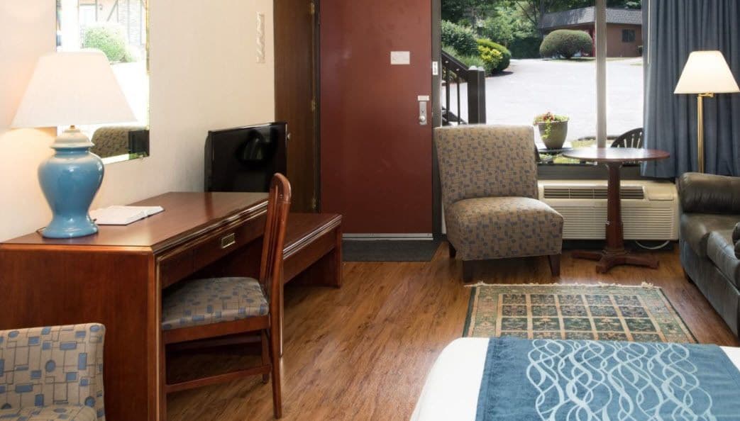 A hotel room with a wooden desk, a patterned armchair, a sofa, and a bed with a blue patterned bedspread.