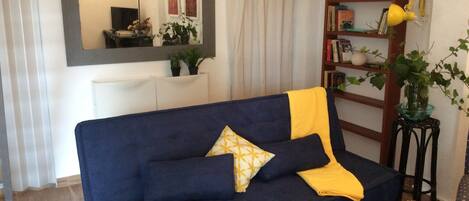 A blue sofa with yellow cushions, a small side table with a plant, and a bookshelf with books and decorative items.
