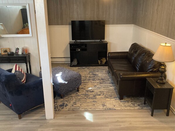 A living room with a dark leather sofa, a small ottoman, a television on a stand, a lamp, and a patterned rug. - Cozy home, quiet neighborhood, near OOB attractions! (Old Orchard Beach)