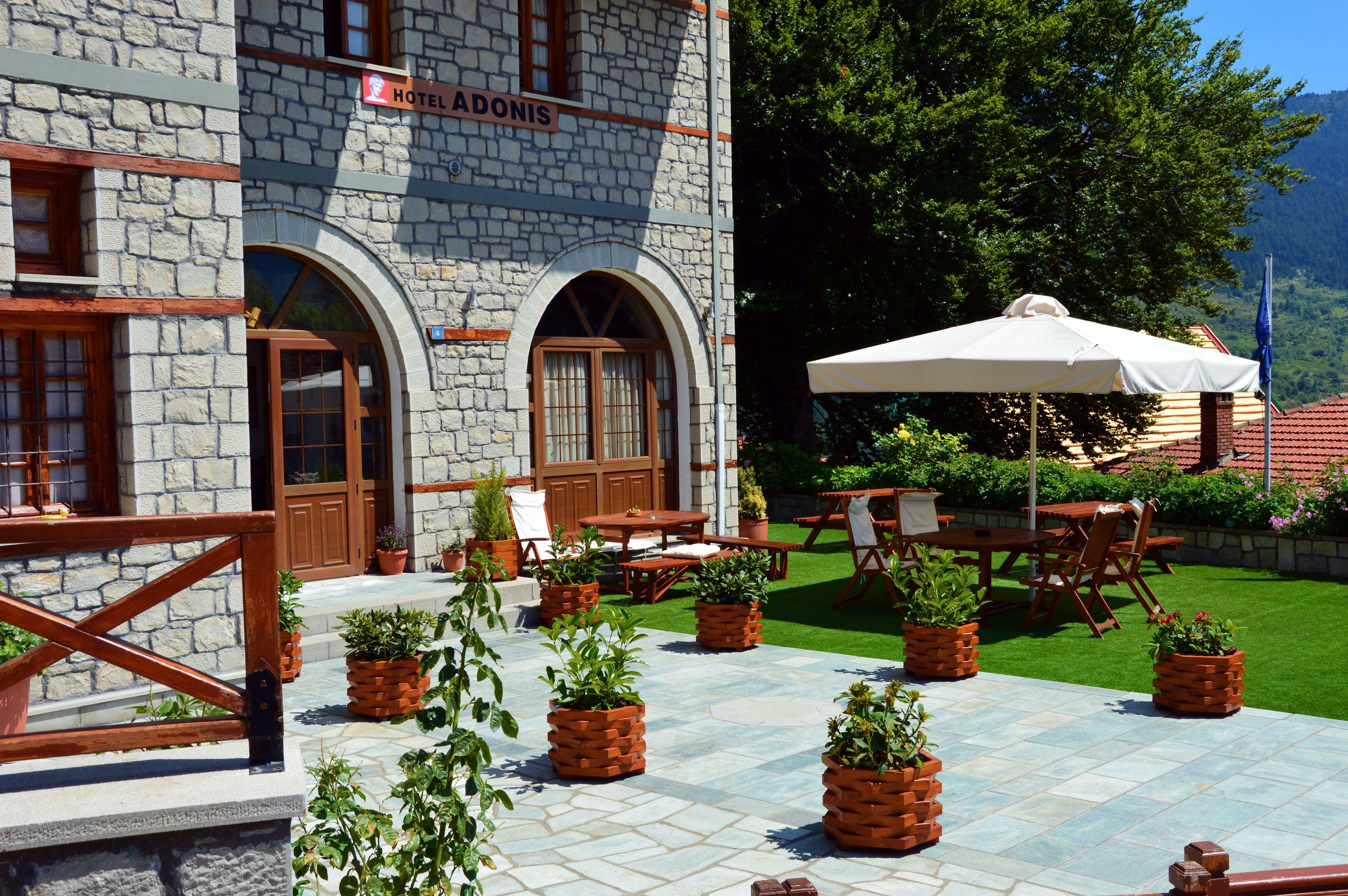 A stone building with a sign reading "HOTEL ADONIS". There are outdoor tables and chairs, and potted plants.