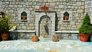 A stone wall with a central arched doorway, flanked by two small windows. There are potted plants on the windowsills and a small tree on the right.