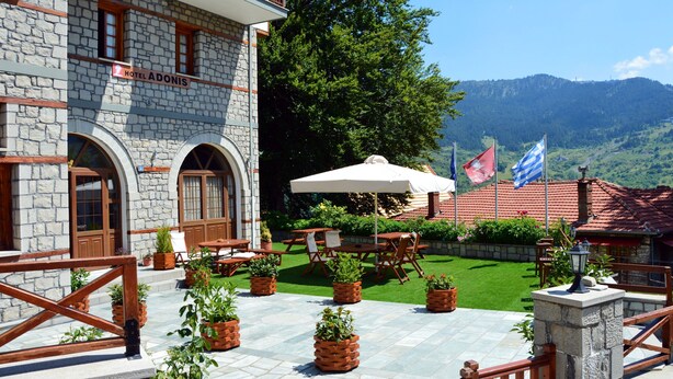 A stone building with a sign reading "Hotel Adonis," outdoor seating with umbrellas, and a view of green hills in the background.
